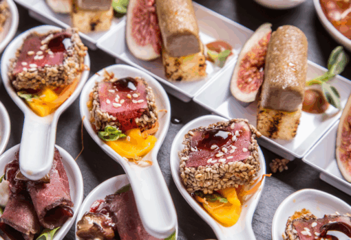 close-up view of a well-organized set of appetizers -including ceviche, roasted meats, fruits, and garnishes- served on white spoons during a lunch coconut grove